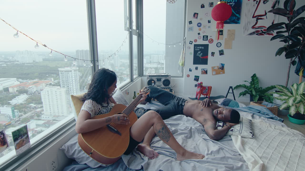 Black Man Listening to Girlfriend Singing and Playing the Guitar in the Bedroom