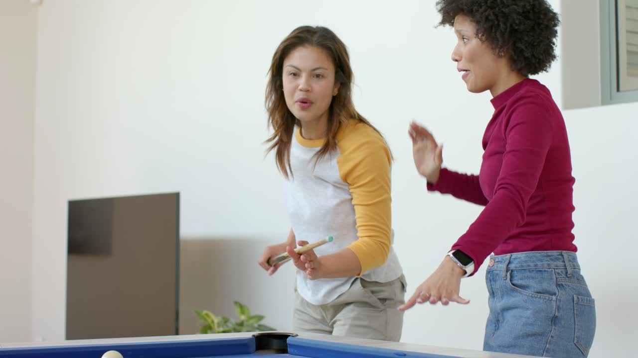 Two female friends playing pool on blue-felt table at home, cue ball striking ball high-fiving