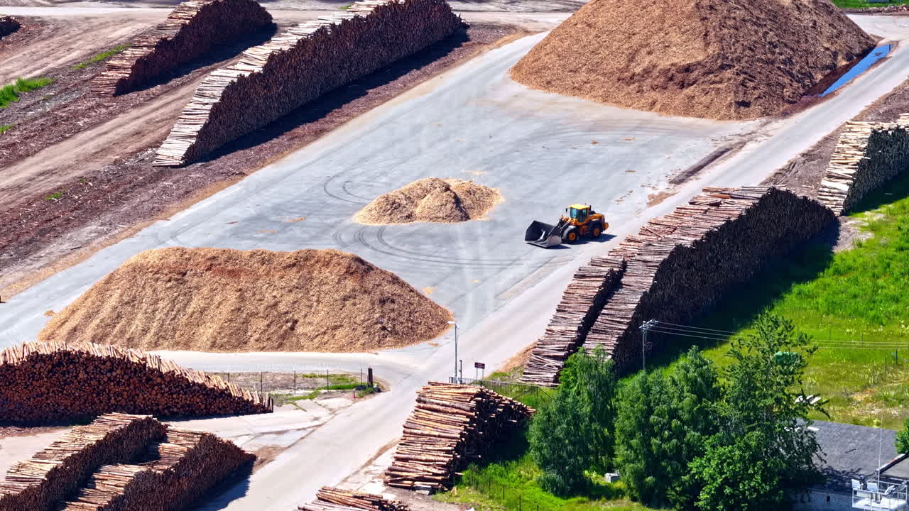 Industrial timber yard at Mērsrags Port, Latvia, showing a front loader working amongst huge stacks of logs and piles of wood chips for processing and export - aerial trucking