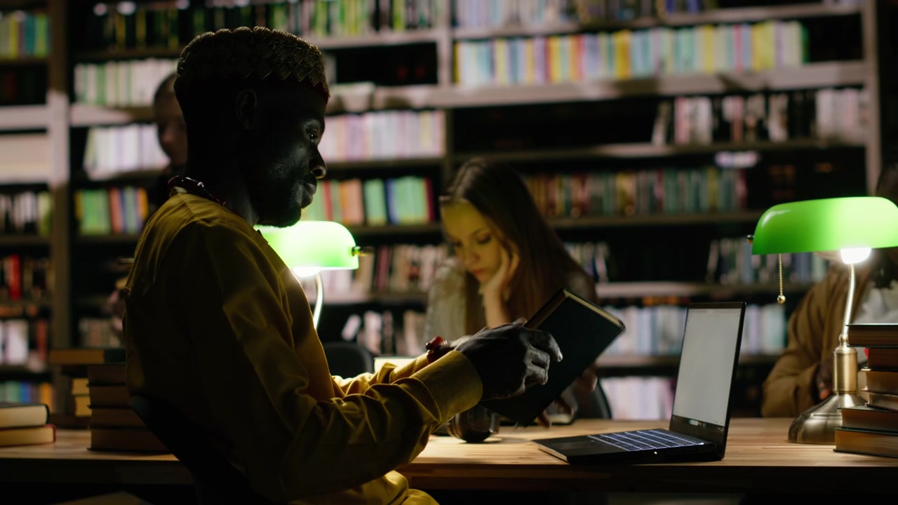 Side view of black man in traditional clothes typing on laptop at library desk
