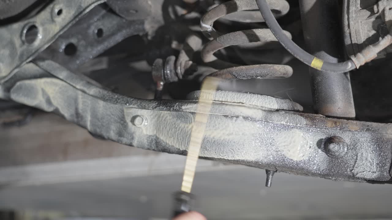 Person applying lanolin rustproofing under car, coating suspension and control arms with pressurized spray gun, close up