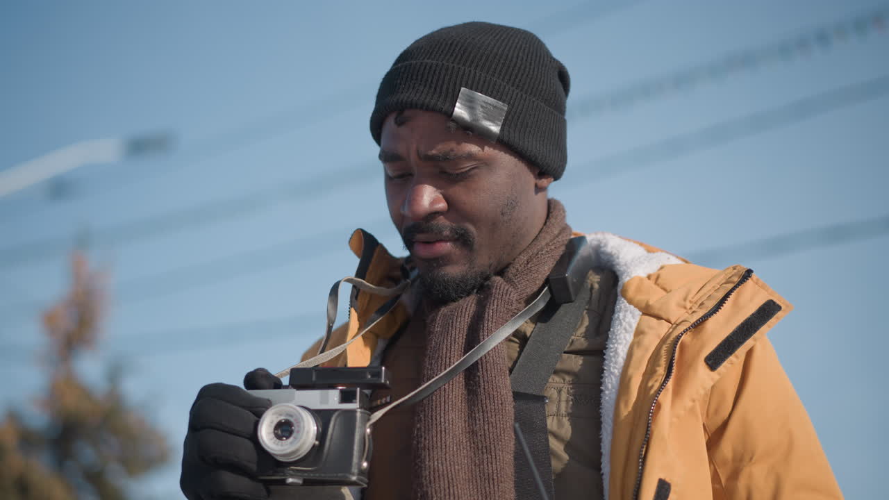 close up sky angle view of freelance photographer holding camera in one hand looking around urban cityscape under bright winter sun wearing gloves beanie jacket capturing candid travel moment