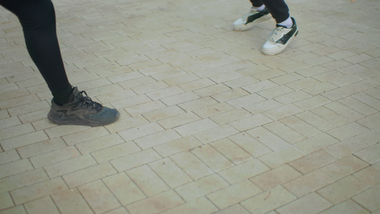 close up feet on brick pavement showing two dancers in playful footwork duel, sneakers tapping and shuffling, dark leggings and white socks visible, urban plaza background, rhythmic motion and casual