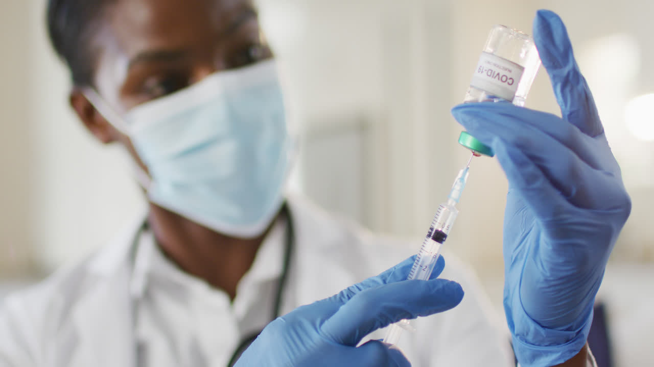 African american female doctor wearing face mask preparing covid vaccine for patient