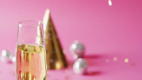 Video-Eines-Vollen-Champagnerglases-Mit-Goldenem-Partyhut-Und-Kugeln-Auf-Rosa-Hintergrund