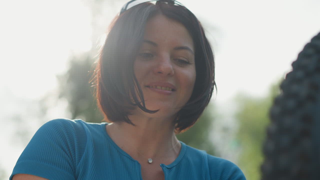 Žena v přírodě se usmívá vedle kola, rekreační cyklistka kontroluje pneumatiku ve sluncem zalitém parku, krátké mikádo jí vlaje kolem obličeje, modrý top, jemné protisvětlo, detailní záběr z nízkého úhlu, klidná a uvolněná nálada, decentní bokeh