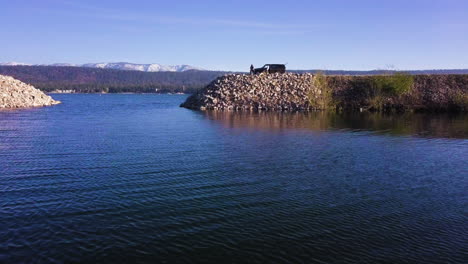 Man-drinking-and-fishing-on-rocky-shore-of-Big-Bear-Lake,-aerial-flying-forward