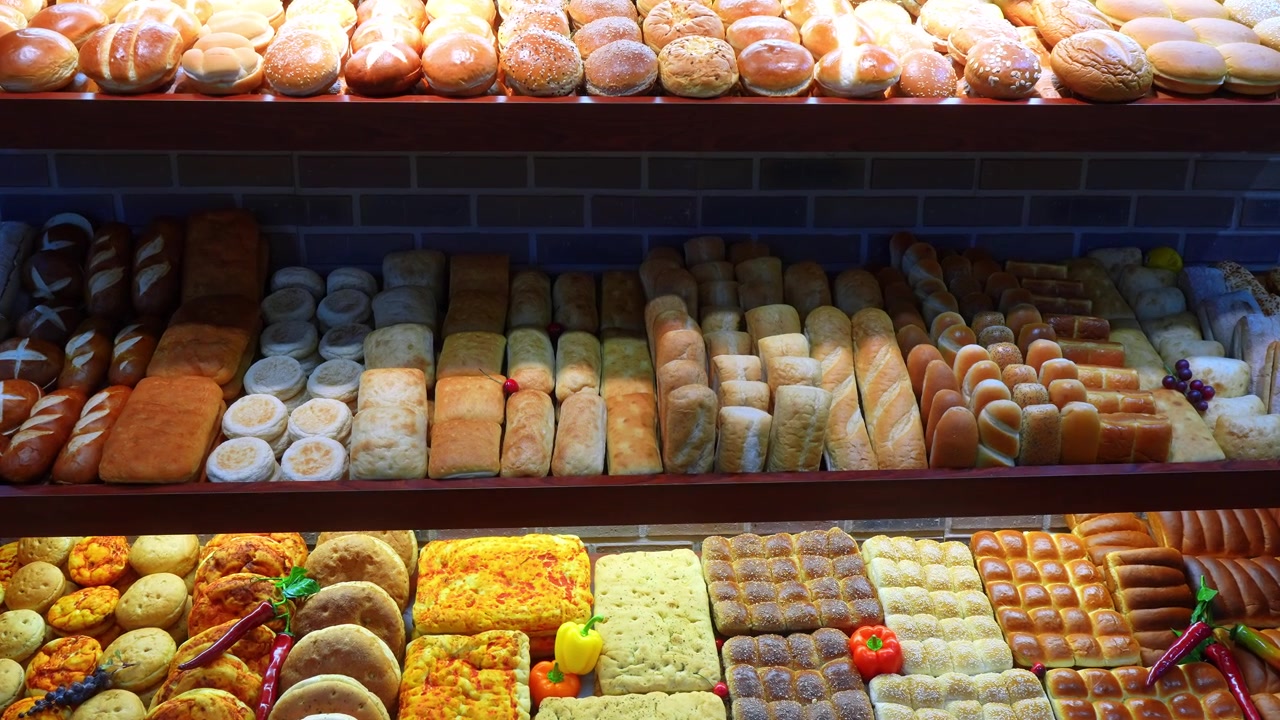 Abundantes filas de diversos panes y bollos en estantes de panadería. Una vista frontal de un expositor de mercado de varios niveles con baguettes, panecillos de hamburguesa y panes planos decorados de colores.