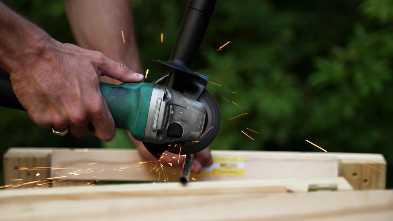 Man using an angle grinder to cut a thin steel pole which is being supported by wooden trestles