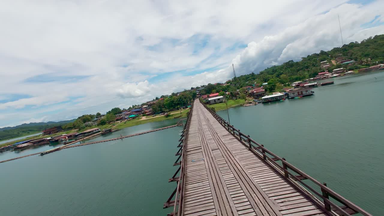 Drone soars above Mon Bridge in Sangkhlaburi, Thailand, revealing the wooden span across the river with floating houses and village scenery surrounded by lush green hills