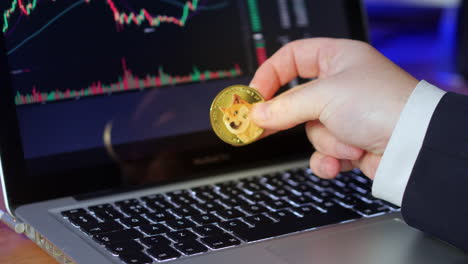 businessman flips over doge coin in his fingers whilst looking at the exchange charts and prices on a laptop