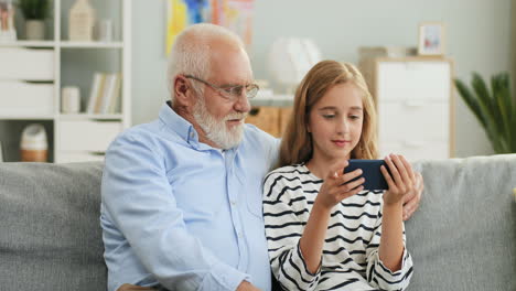 Portrait-Shot-Of-The-Grandfather-And-Granddaughter-Resting-On-The-Couch-In-The-Living-Room-And-Girl-Showing-Something-To-The-Grandpa-On-The-Phone-Screen