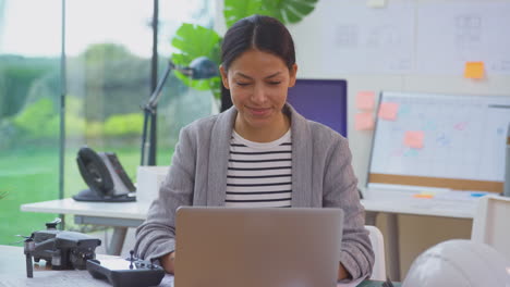 Architektin-Im-Büro-Am-Schreibtisch-Mit-Drohne,-Die-Mit-Laptop-An-Plänen-Für-Ein-Neues-Gebäude-Arbeitet