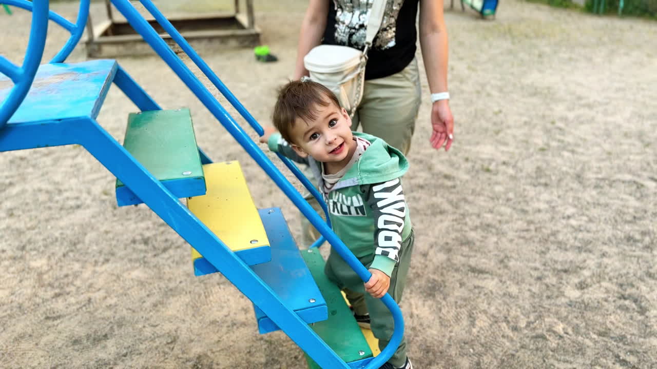 Happy active toddler going up by the stairs of the slide. Caring mom securing her baby. Playground backdrop.