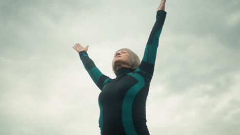 woman standing under cloudy sky practicing yoga, lifting arms in a wide stretch, focusing on slow and controlled movements for balance and relaxation, she is dressed in a green and black yoga suit