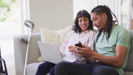 Happy-biracial-couple-in-living-room-using-laptop-and-smartphone,-with-crutch-leaning-on-couch