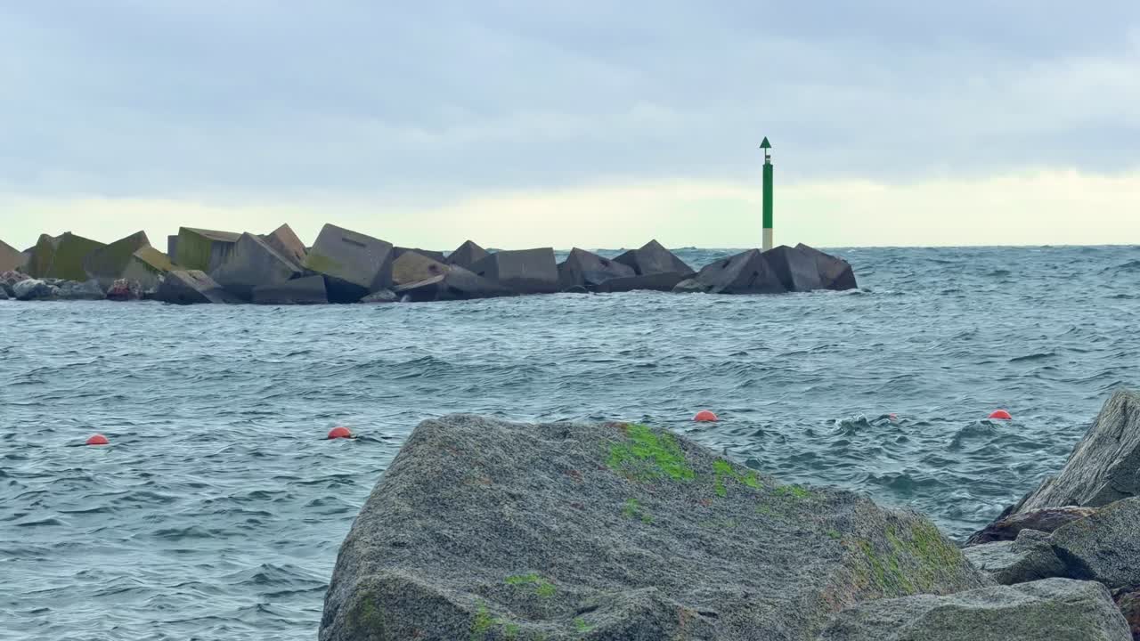 Prise de vue statique d'un brise-lames et d'une balise de navigation verte sur le littoral de Barcelone, avec des vagues, un rocher au premier plan et la mer Méditerranée nuageuse. Espagne