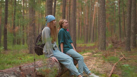 two tired hikers rest on fallen tree in serene forest, one with blue bandana lifting leg happily and looking relaxed, while companion in green shirt places hand on tree for support