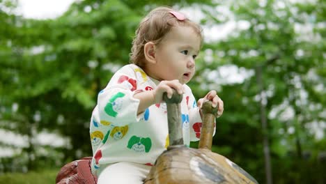 Adorable-Bebé-Asiático-Multiétnico-Montando-Un-Bicho-Mecedor-De-Madera-En-El-Parque-De-La-Ciudad