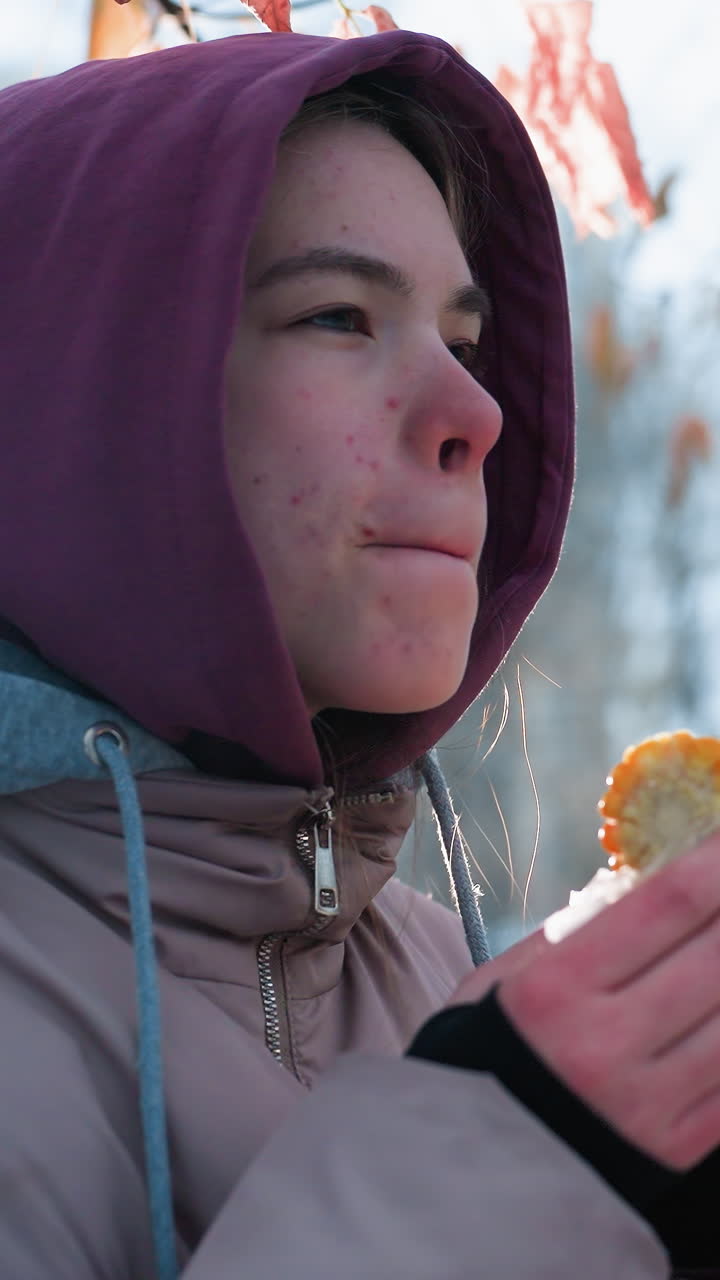 vista lateral de una mujer comiendo maíz con hojas secas balanceándose suavemente por encima, fondo borroso con personas caminando en el parque durante el invierno, luz natural suave reflejándose en su cara, momento pacífico del parque