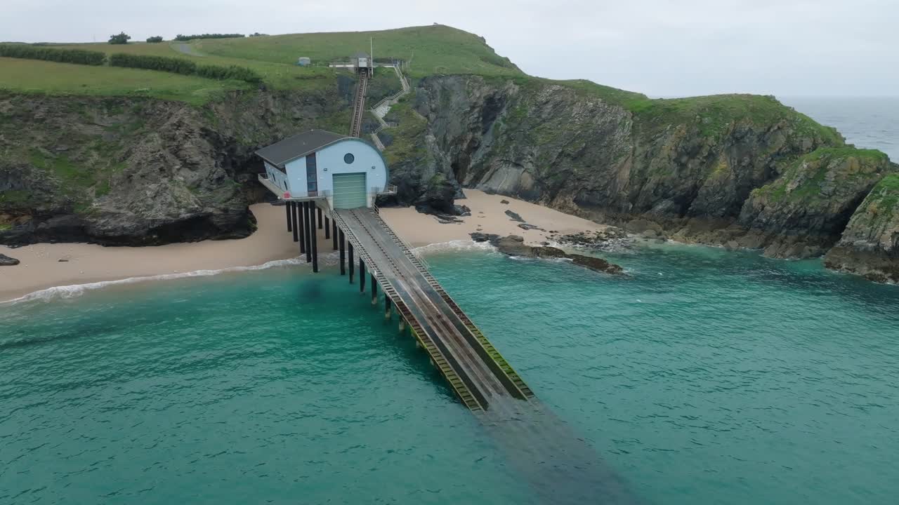 RNLI lifeboat station with long slipway descending into clear blue water next to jagged cliffs and sandy beach. Camera descending orbit. Mother Ivey's Bay, Cornwall, UK.