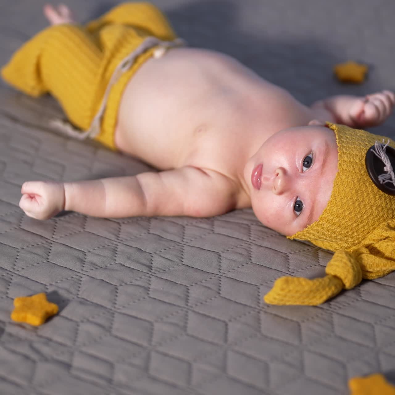 Cute baby boy in a yellow costume of a gnome lies on the bed. Decorative stars around the baby on the bed. Baby sticks out tongue