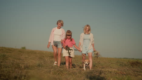 two women walking alongside little child holding dog leash while dog pauses to look up at woman in pink, woman in blue holds bouquet of flowers, scene conveys family connection