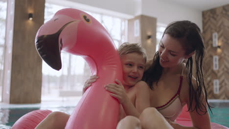 happy cute boy is sitting on inflatable flamingo with mother in swimming pool family center for rest and have fun