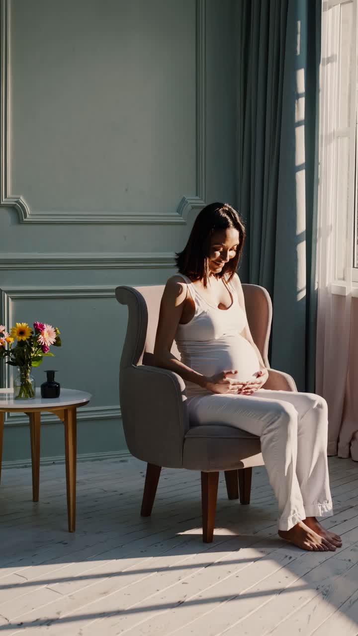 A serene video scene of a pregnant woman in a sunlit room, sitting in a chair, captured from a side