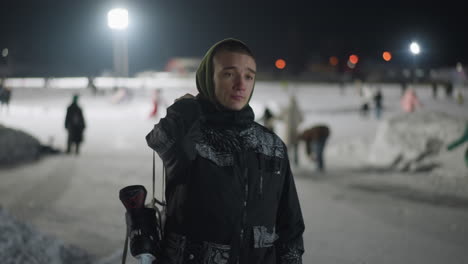 young man in patterned winter jacket walks confidently on ice rink at night holding skate boot over shoulder with blurred background of skaters and glowing arena lights under cold winter sky