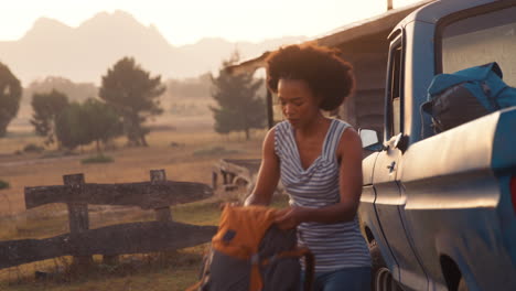 Portrait-Of-Woman-Unloading-Backpacks-From-Pick-Up-Truck-On-Road-Trip-To-Cabin-In-Countryside