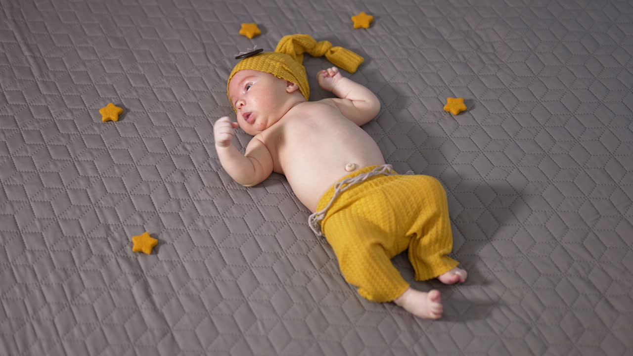 Adorable kid wearing yellow hat and pants lying on the bed. Little boy tossing his little hands and feet. Baby on the grey background view from above.