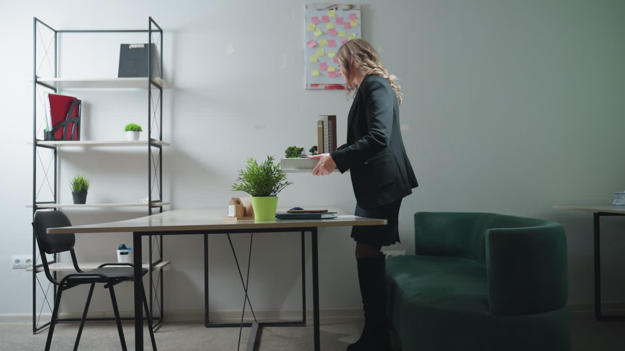 Real estate agent carrying building model walks toward desk and drops it in workplace with minimalist decor, featuring desk plant, bookshelves, green couch and modern layout