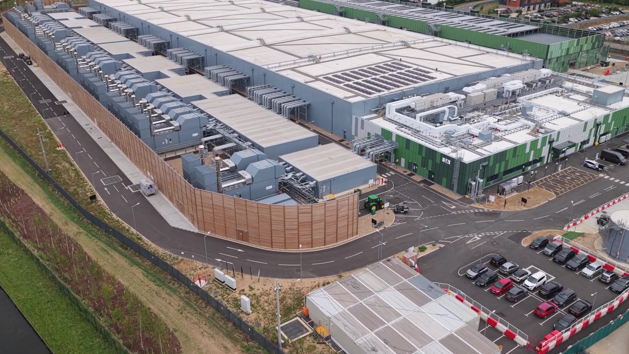 Moderne serverkompleks infrastruktur viser kølesystemer og byggeaktivitet i dagslys nær en kanal med aktive transportkøretøjer, der bevæger sig rundt på stedet i eftermiddagstimerne