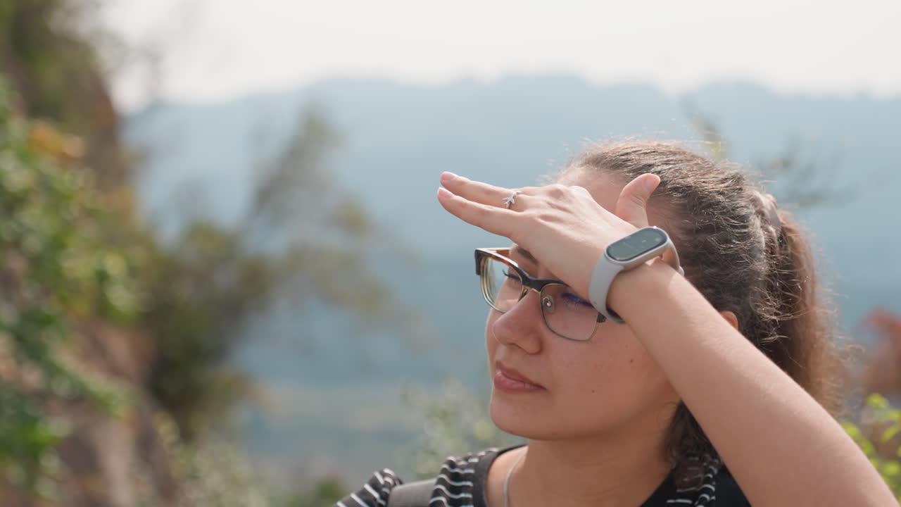 Woman tourist shading eyes at lookout, hand over brow, eyeglasses and smartwatch visible, distant ridgelines and valley, soft sunlight and contemplative expression during short rest