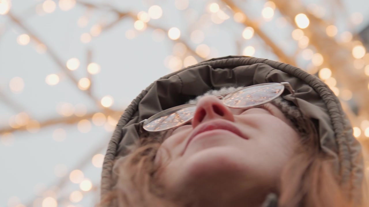 Žena hledící na blikající světla, tichá noční snílka zastavující se na prázdné ulici, třpytivé bokeh větve nahoře, měkký kabát a ozdobné brýle, introspektivní nostalgická atmosféra.