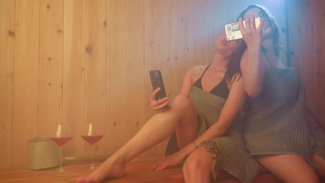 two women wrapped in towels sit close on wooden sauna bench, steam drifting as they smile and pose for selfie with phones in hand, drinks nearby creating cheerful relaxed atmosphere of friendship