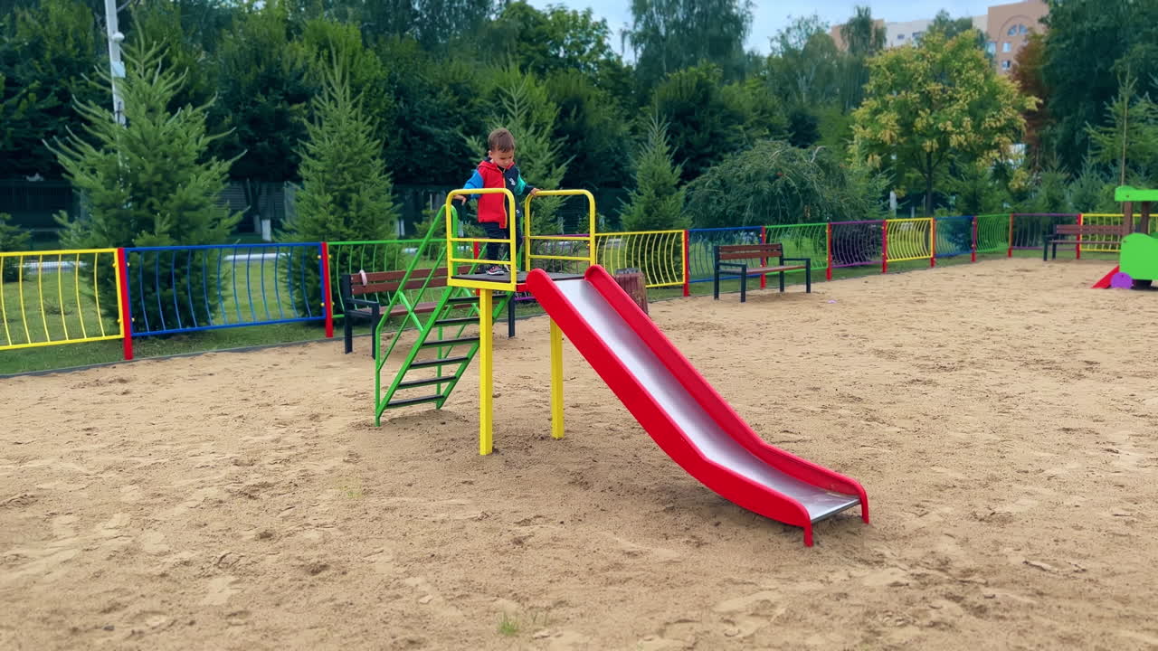 Lovely kid goes up by the stairs on the slide. Toddler boy sits on top, taps his feet and goes down.