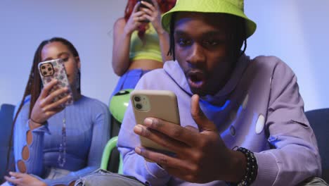 Close-Up-Studio-Shot-Of-Young-Gen-Z-Friends-Sitting-On-Sofa-Sharing-Social-Media-Post-On-Mobile-Phones-1