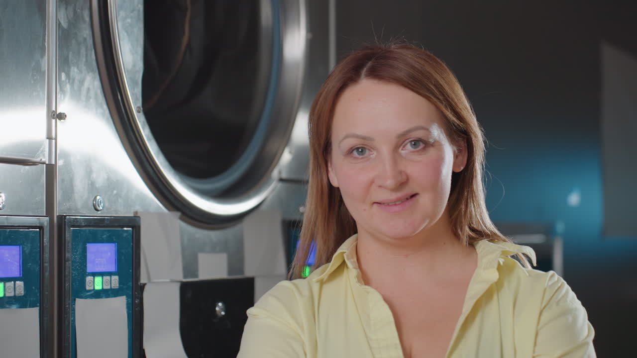 Portrait of work woman posing near industrial washing machine inside laundromat, confident employee in yellow shirt looking aside, stainless drum and control screens in background