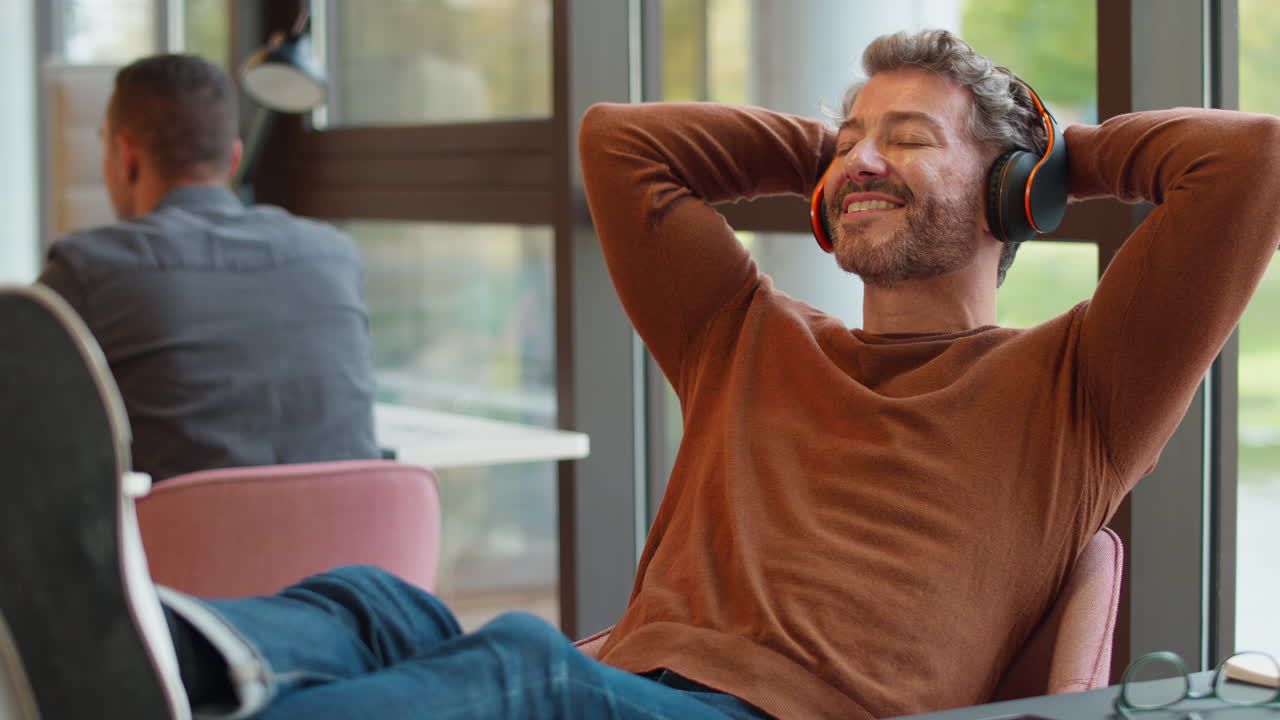 Mature Businessman With Feet On Desk In Office Listens To Music On Wireless Headphones