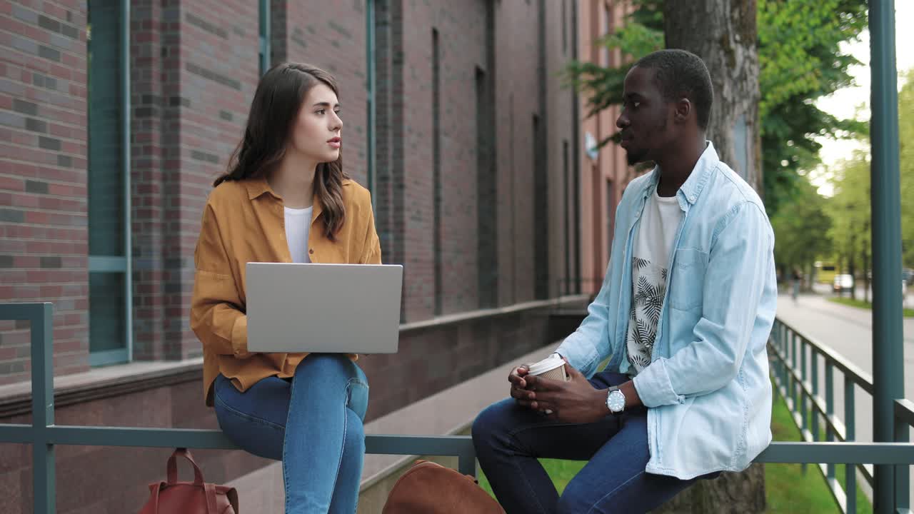 Caucasian and african american students talking while drinking coffee and using the laptop sitting in the street near the college