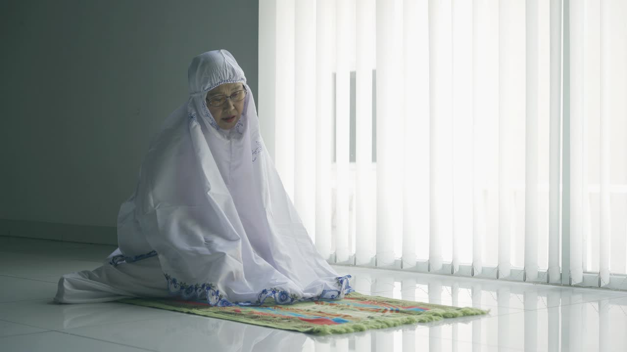 시니어 무슬림 여성이 창문 근처에서 혼자 기도하고 있습니다.