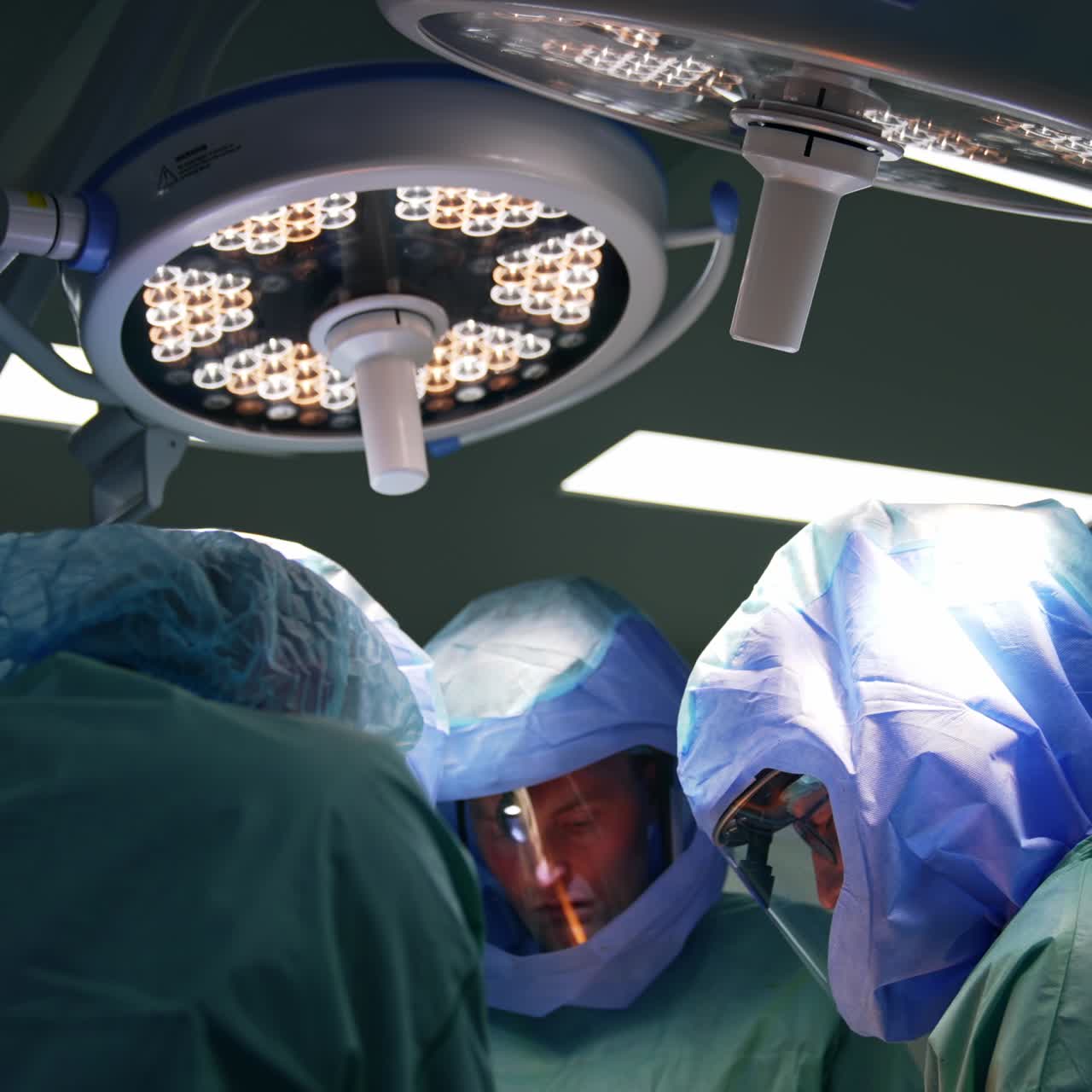 Medics bent over the patient in modern surgery room. Doctors in protective suits work under the round lamps in operational theatre