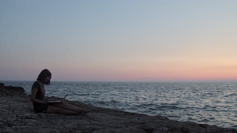 Girl-sitting-by-the-sea-with-laptop-blue-hour,-waves-hitting-rocks,-slow-motion