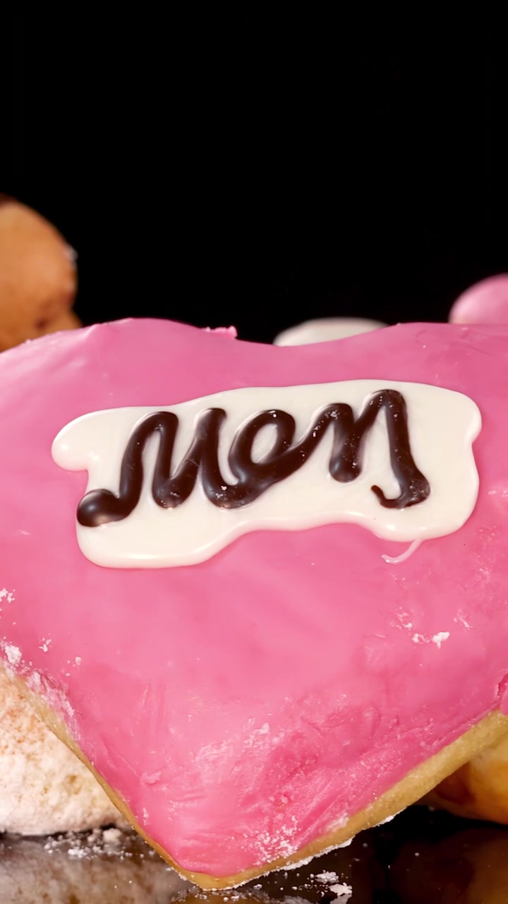 明るいスタジオ照明の下、黒い背景をバックに、ピンクのアイシングとチョコレートで「Mom」と書かれたハート型のドーナツが、様々なドーナツの前で回転している。