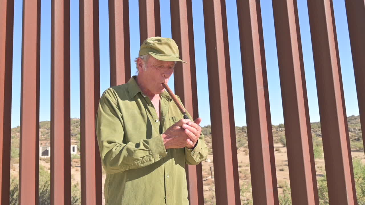 Older man in front of US Mexico border fence lights a cigar.