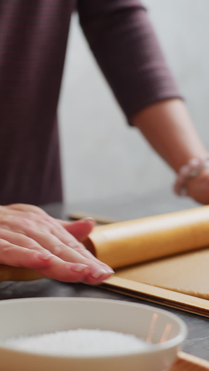 Baker hands rolling brown dough with wooden rolling pin, left wrist adorned with pink beaded bracelet, slight blur in background featuring candle, kitchen tools