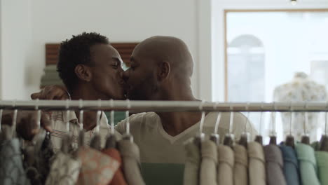 primer plano de una pareja gay negra besándose detrás de un estante de ropa en la tienda.