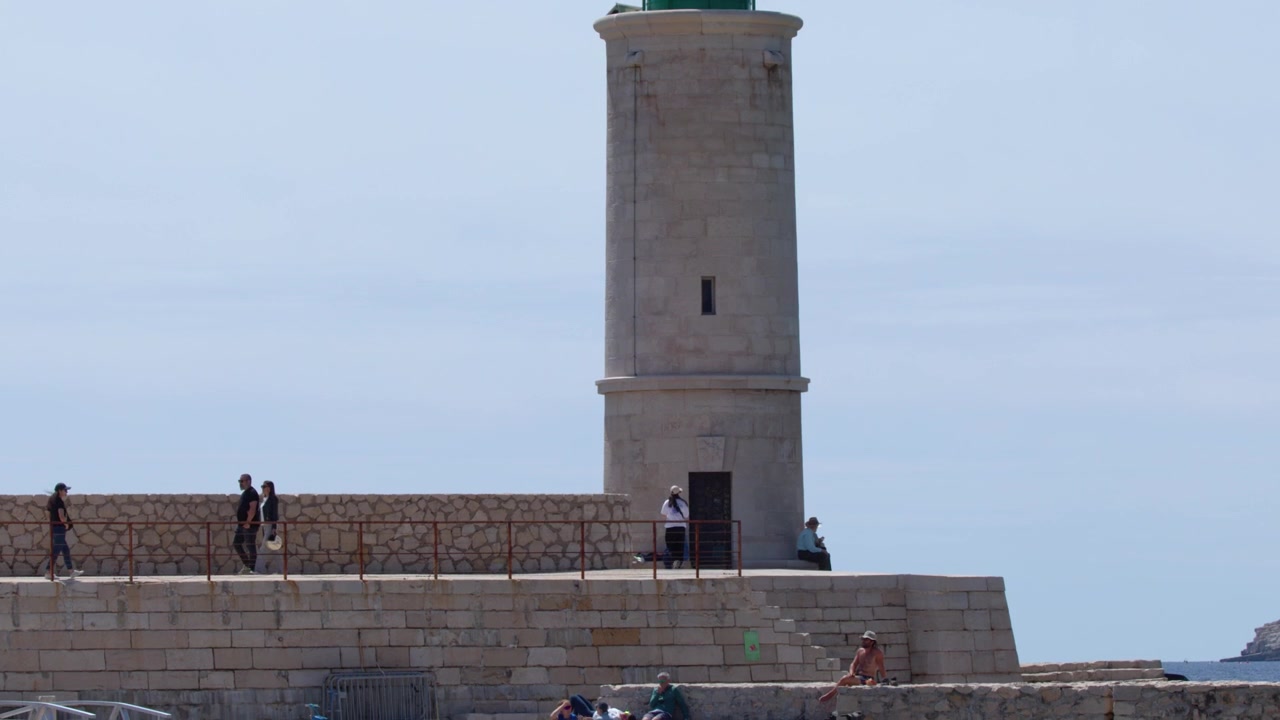 Inquadratura media statica di un faro in pietra e un molo con persone che camminano a Cassis, Francia.